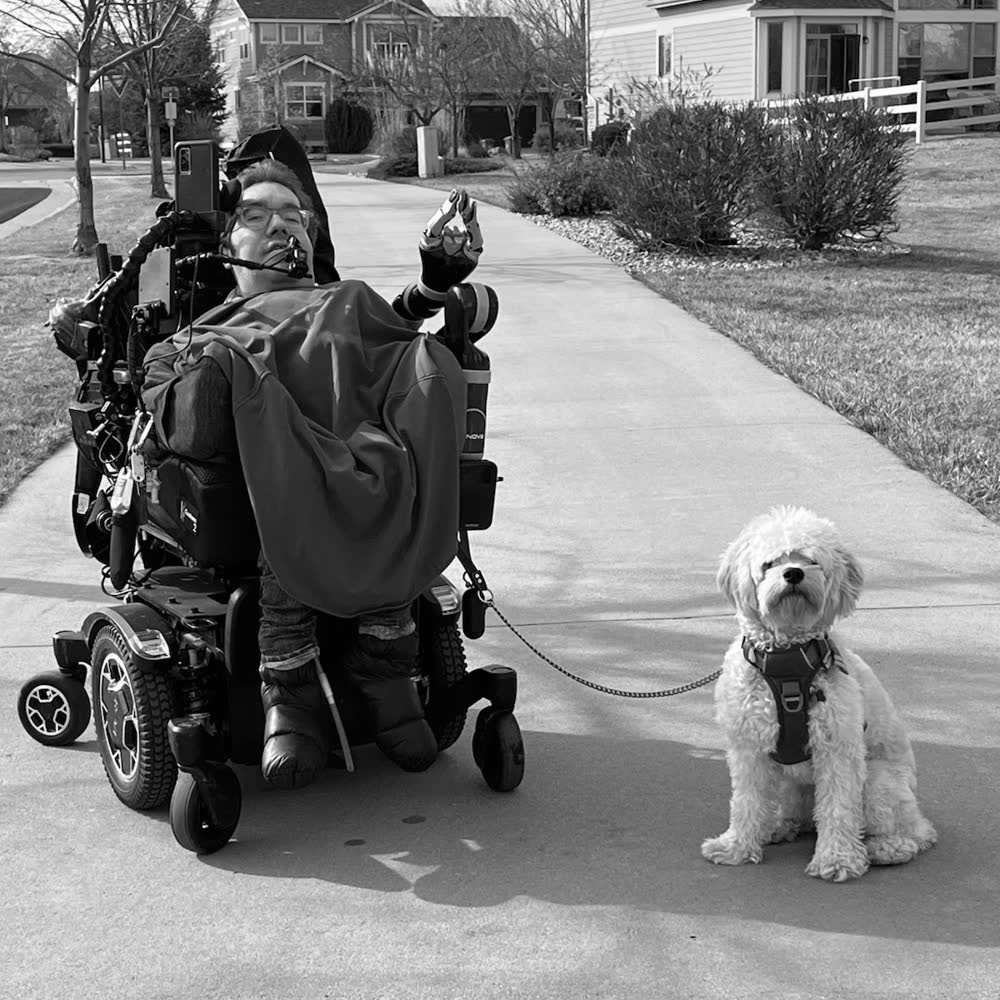 Owen outdoors on a suburban footpath in his powered wheelchair, with Buddy, a small cream-colored curly-haired dog, sitting on a lead beside him.